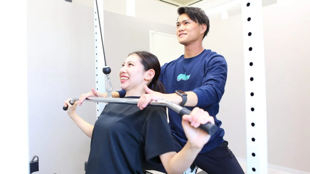 ラットプルダウンをする女性の肘を支えて指導するトレーナー
