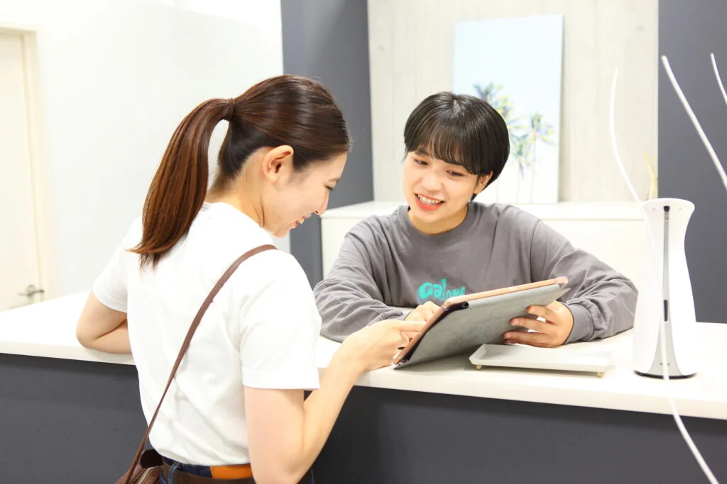 受付カウンターでタブレットを見せながら女性に案内するスタッフ