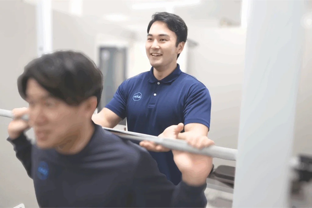 バーベルを担ぐ男性の横で笑顔でサポートするトレーナー